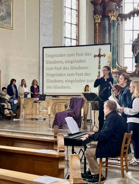 Menschen mit und ohne Behinderung im Altarraum, die singen. Eine Band macht Musik und eine Gebärdensprachdolmetscherin übersetzt den Liedtext. Dieser wird auf einer Leinwand angezeigt.